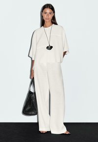 Une femme se tient dans une tenue blanche ample et légère avec un pantalon large, tenant un sac tissé noir et portant un collier avec un pendentif noir, le tout sur un fond clair.