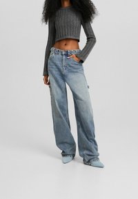 Person wearing a gray cropped ribbed sweater, loose light blue high-waisted jeans, and blue pointed-toe shoes, standing with one hand in pocket.