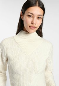 Cream-colored knit sweater with a high collar, featuring a textured zigzag pattern and ribbed detailing on the collar.