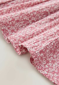 Różowa tkanina w kwiatowy wzór z białymi kwiatami, o teksturowanej powierzchni i plisowanym wzorze. Krawędzie są miękkie i postrzępione, co tworzy delikatny wygląd.