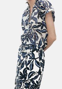 Personne portant une tenue blanche avec des motifs de feuilles noires et bleues, à manches courtes, et une jupe à taille nouée, debout devant un fond uni.