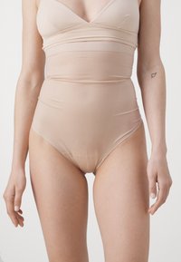 Beige body gemaakt van een gladde, rekbare stof. Beschikt over een diepe halslijn, hoge uitsnijdingen en een naadloos ontwerp met ribbelstructuuraccenten.