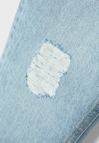 Tessuto di denim blu chiaro con un'imponente toppa strappata dai bordi sfrangiati, posizionata su uno sfondo testurizzato dello stesso materiale.