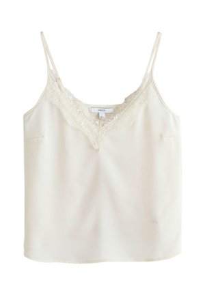 Femme aux cheveux bruns courts portant un caraco blanc orné de dentelle et un pantalon marron, debout contre un mur lambrissé en bois.
