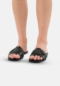 Sorte slidesandaler med krydsede stropper lavet af glat materiale, firkantet tådesign, flad sål og synlige tånegle med lys neglelak.