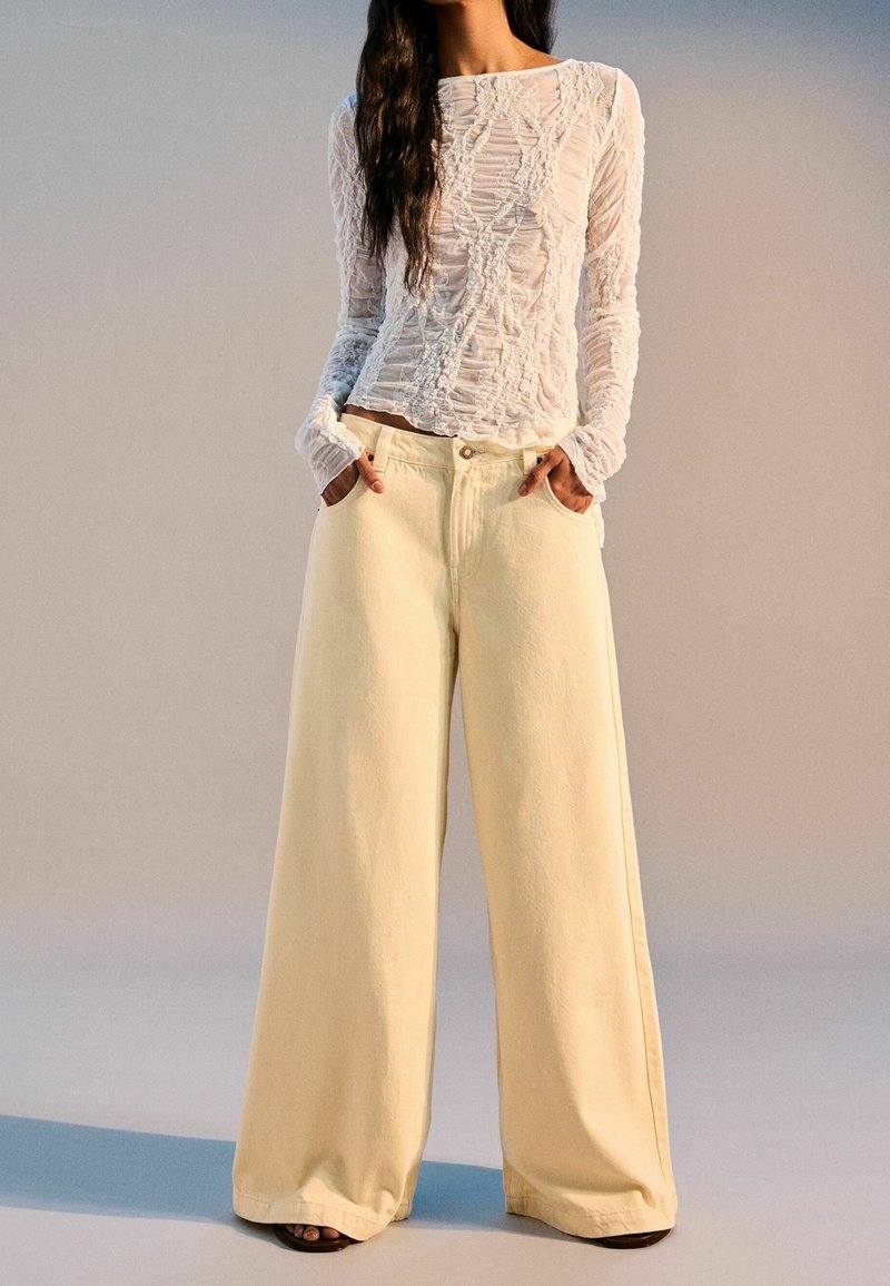 Top blanco de manga larga con textura y detalles recogidos, combinado con pantalones de pierna ancha de un suave color amarillo pálido, ambos confeccionados con materiales suaves.