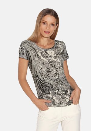 T-shirt noir et blanc à motif paisley avec des manches courtes, un col rond et un tissu lisse et extensible. Le modèle pose les mains dans les poches.