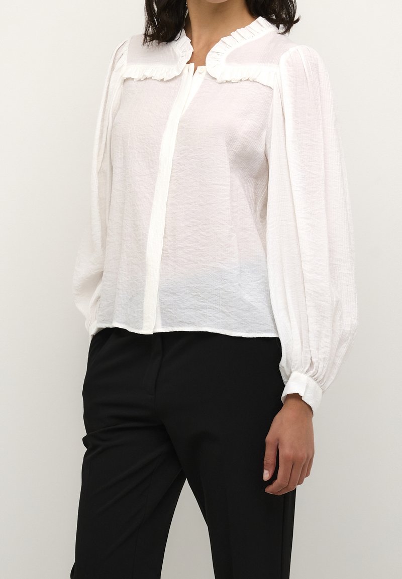 White, textured blouse with a ruffled collar, button-up front, and long, puffed sleeves. Paired with black trousers.