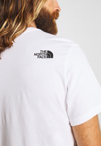 Man med långt hår och skägg iförd en vit T-shirt med The North Face-loggan på övre ryggen mot en enhetlig bakgrund.