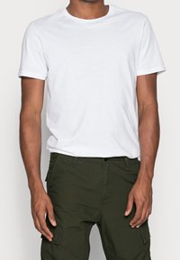 Camiseta blanca de manga corta con cuello redondo, fabricada en algodón, con una textura suave y un corte clásico, combinada con pantalones cargo de color verde oscuro.