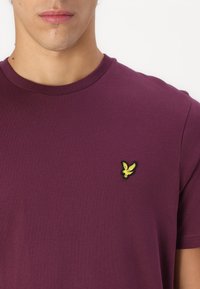 T-shirt violet à manches courtes et col rond en coton, avec un petit logo d'oiseau brodé jaune sur la partie gauche de la poitrine.