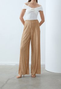 Haut blanc léger à épaules dénudées avec des détails froncés et une taille accentuée en or, associé à un pantalon doré large, à plis et à taille haute.