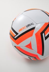Balts un oranžs sintētiskais futbola bumbas ar melniem akcentiem, kas izceļas ar leņķainiem rakstiem un redzamu "Mercurial" logotipu.