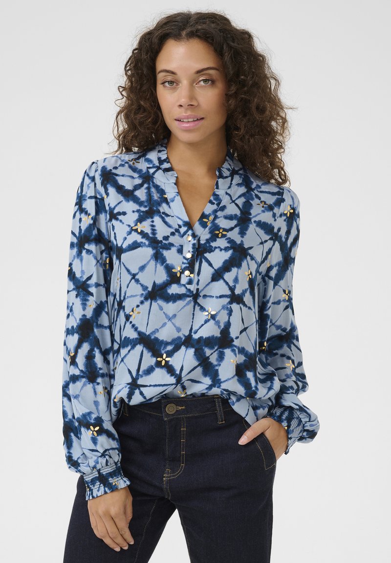 Chemise bleu tie-dye avec une coupe décontractée, des manches longues, une patte de boutonnage et des accents floraux dorés. Le tissu semble léger avec une texture douce.