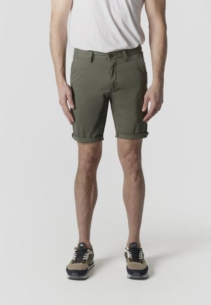 Férfi, aki felhajtott olívazöld rövidnadrágot, fehér ujjatlan inget és bézs-fekete sportcipőt visel, sík világos háttér előtt áll.