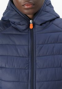 Nahaufnahme einer Person, die eine marineblaue Steppjacke mit Kapuze und einem zentralen Reißverschluss trägt, der einen orangefarbenen Zipper hat.