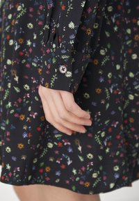 Abito in tessuto nero con motivi floreali colorati. Presenta polsini con bottoni e chiusura a cerchio. Texture morbida e vestibilità comoda.