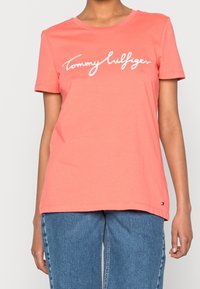 Osoba w koralowej koszulce z krótkim rękawem i białym napisem "Tommy Hilfiger", w niebieskich dżinsach, stojąca na tle jasnego, jednolitego tła.