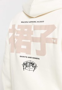 Felpa crema con grande testo giapponese rosa morbido e logo nero "SAINTS AND SINNERS". Il materiale è liscio e presenta una vestibilità comoda.