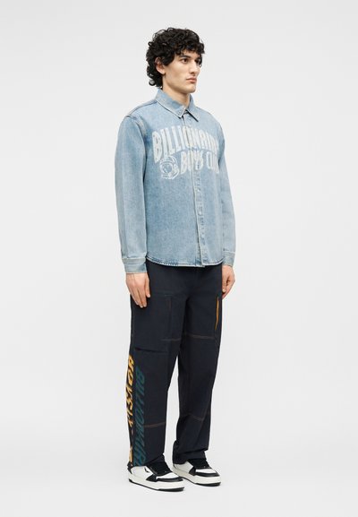 Άνδρας με σγουρά μαλλιά που φοράει ανοιχτό μπλε τζιν πουκάμισο με στάμπα "Billionaire Boys Club", μαύρο παντελόνι cargo με κείμενο στο πλάι και μαύρα-λευκά αθλητικά παπούτσια.
