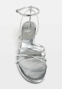 Sandale argentée à talons hauts avec une finition métallique, comportant plusieurs fines lanières et une lanière de cheville. Texture lisse et design à bout ouvert.