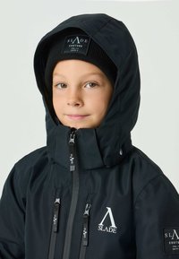 Black hooded jacket with zipper closures and logo accents. Features a soft, smooth texture and is paired with a black knit beanie.