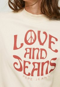 T-shirt color crema con un audace design grafico rosso che presenta il testo "LOVE AND JEANS" e un simbolo di pace. Tessuto morbido.