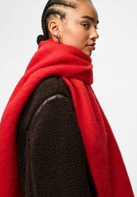 Sciarpa di lana rossa avvolta attorno al collo, stratificata su un giubbotto marrone con texture e un accento in pelle liscia.