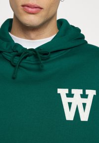 Zielona bluza z kapturem, wykonana z miękkiego materiału, z kieszenią typu kangur, sznurkiem w kapturze oraz dużą białą haftowaną literą "W" na klatce piersiowej.