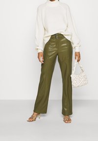 Maglione crema con texture a coste e collo alto. Pantaloni in ecopelle verde oliva con cuciture frontali. Borsa bianca intrecciata e sandali con tacco dorati.