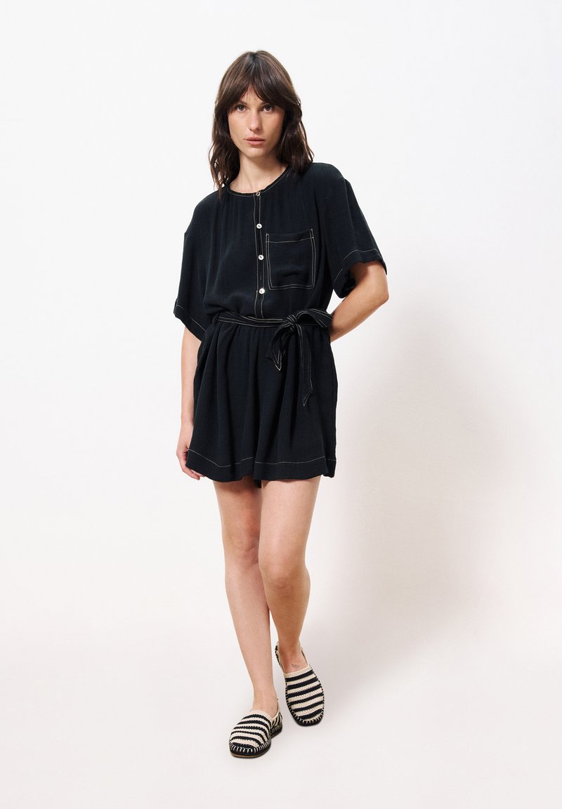 Schwarzes Kurzarmkleid mit Tasche, Knopfdetails und tailliertem Schnitt. Kombiniert mit gestreiften Slippern. Glatte Stofftextur.