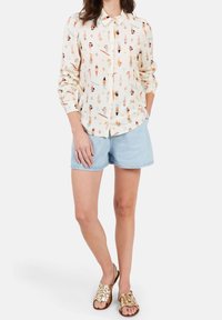 Chemise boutonnée crème avec un motif ludique de personnages illustrés, associée à un short en jean bleu clair et des sandales tissées dorées.