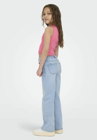 Jean flare bleu clair, en denim, taille mi-haute, poches arrière visibles. Associé à un haut sans manches rose et des baskets à plateforme blanches.