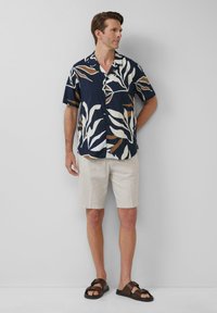 Marineblaues Kurzarmhemd mit weißem und braunem Blattmuster, kombiniert mit beigen Shorts und braunen Sandalen. Glatte Stoffbeschaffenheit, lässige Passform.