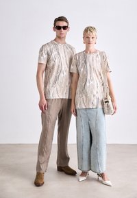 To modeller har matchende beige mønstrede t-shirts, der er parret med beige bukser og en lang denimnederdel. Tilbehøret inkluderer solbriller og en lille taske.