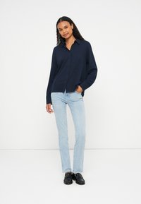 Femme aux cheveux bouclés portant une chemise boutonnée bleu marine, un jean bleu clair et des ballerines noires, se tenant contre un fond blanc uni.
