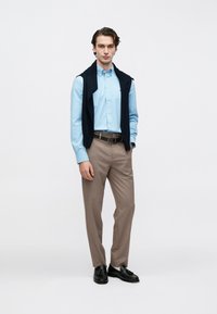 Chemise bleue claire boutonnée, pull bleu marine drapé sur les épaules, pantalon beige, chaussures noires, coupe ajustée, tissu lisse, design minimaliste.
