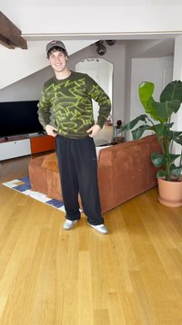 Zielony sweter z abstrakcyjnymi żółtymi wzorami, czarne spodnie i srebrne buty; stoi w pokoju z drewnianą podłogą, rośliną i kanapą.