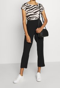 Kvinna i top med zebramönster, högmidjade svarta cropped byxor, vita sneakers och håller en svart crossover-väska.