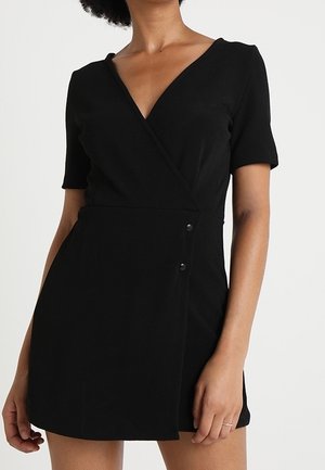 Femme portant une robe portefeuille noire courte à manches courtes avec deux boutons visibles sur le côté, sur un fond uni.