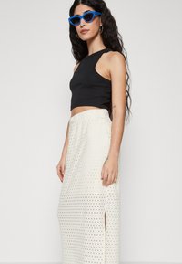 Sort crop-top med høj hals; hvid maxiskirt med perforeret mønster og sideslids; blå katteøjne solbriller.