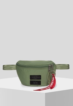 Sac banane vert avec des patchs noirs et deux curseurs de fermeture éclair, dont un accroché à une étiquette rouge avec du texte blanc, exposé sur un socle blanc.