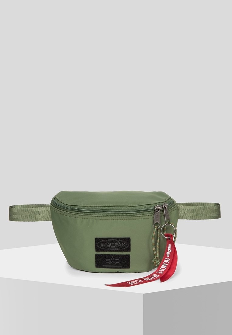 Sac banane vert avec des patchs noirs et deux curseurs de fermeture éclair, dont un accroché à une étiquette rouge avec du texte blanc, exposé sur un socle blanc.
