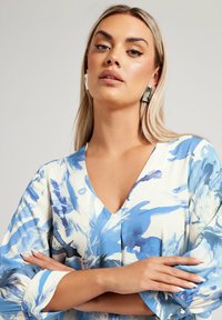 Lätt, blå blommönstrad blus med V-ringning och flödande ärmar. Slät tyg, stora abstrakta blå och vita penseldrag.