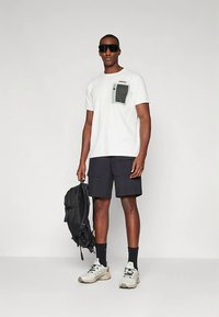 Vīrietis baltā T-kreklā ar melnu kabatu, melnās kravas šortos, sporta apavos, melnās zeķēs, lielās saulesbrillēs, tur melnu mugursomu pie sāniem.