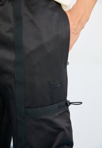 Calças atléticas pretas feitas de um tecido liso com um cós elástico, apresentando um discreto detalhe de 3 riscas e um pequeno logótipo em relevo.