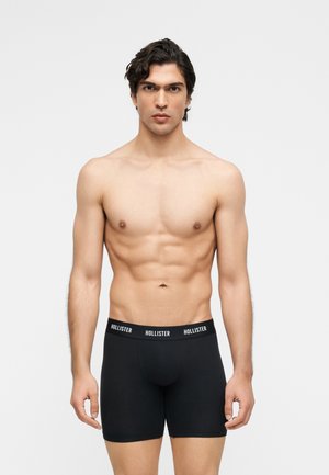 Mladý muž s tmavými vlasmi, oblečený v čiernych boxerkách Hollister, stojaci pred jednoduchým svetlosivým pozadím.