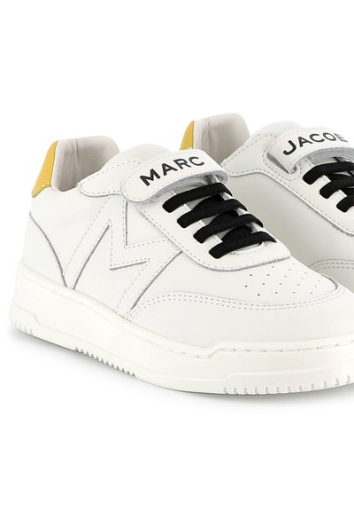 Sneaker in pelle bianca con lacci neri e una fascetta in velcro. Presentano un accento giallo sul tallone e un logo in rilievo sulla linguetta.