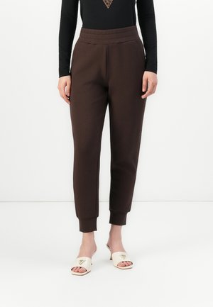 Vrouw in bruine tapers joggers met zijzakken, gecombineerd met witte open teen sleehakken, op een witte achtergrond.