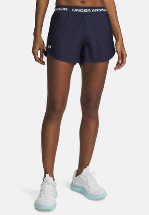 Kvinde iført mørkeblå Under Armour løbeshorts og hvide træningssko med lyseblå såler, stående på en hvid baggrund.